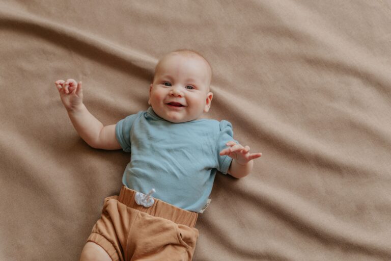 Cute baby lying on a soft beige blanket, smiling and wearing a blue shirt and brown shorts.